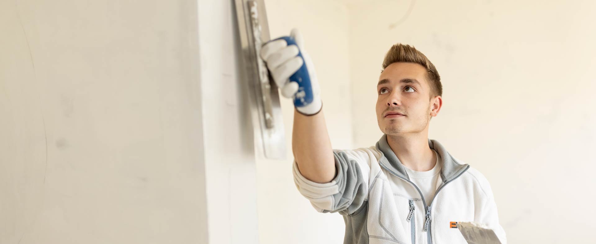 Junger Mann mit weiß-grauer Arbeitsjacke und Handschuhen trägt eine Glättkelle und einen Spachtel und bearbeitet eine Wand