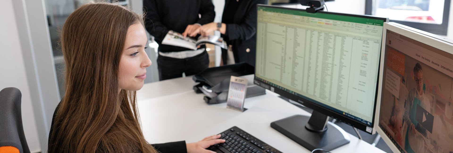Junge Frau sitzt am Schreibtisch vor zwei Monitoren und tippt auf der Tastatur, im Hintergrund stehen zwei Personen und schauen in ein Buch