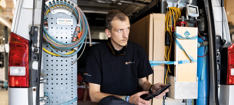 Mann in Arbeitskleidung sitzt im geöffneten Heck eines Transporters und hält ein Tablet in der Hand